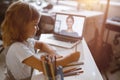 Schoolgirl writes listening to teacher at videocall via modern laptop at table in light room Royalty Free Stock Photo