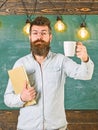 Scientist holds book and drinks coffee, chalkboard on background, copy space. Coffee break concept. Man with beard on Royalty Free Stock Photo