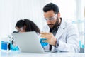 Scientist in lab coat and safety glasses holding flask with blue solution at modern laboratory. Take note on the laptop for Royalty Free Stock Photo