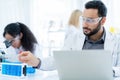 Scientist in lab coat and safety glasses holding test tube with blue solution at modern laboratory. Take note on the laptop for Royalty Free Stock Photo