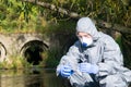 A scientist in a protective suit, mask and gloves, holds in his hand a flask with a blue liquid and takes water from the river for Royalty Free Stock Photo