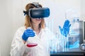 Scientist in VR googles, lab coat and flask flipping through a page of virtual interface with graphs Royalty Free Stock Photo