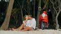 Scooter road trip. Lovely couple on red motorbike in white clothes on sand beach. People walking near the tropical palm Royalty Free Stock Photo
