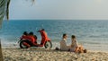 Scooter road trip. Lovely couple on red motorbike in white clothes on sand beach. People walking near the tropical palm Royalty Free Stock Photo