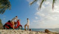 Scooter road trip. Lovely couple on red motorbike in white clothes on sand beach. People walking near the tropical palm Royalty Free Stock Photo