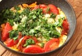 Scrambled eggs with tomatoes, sprinkled with chopped dill and parsley in a black skillet on a brown wooden table.