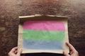 On the scratched table lies an old, tattered piece of paper with a picture of the Polysexual pride flag