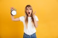 Screaming shocked young blonde woman posing isolated over yellow wall background dressed in white casual t-shirt holding alarm