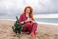 Sea Lady in plaid shirt with a mug in her hands enjoys beach with Christmas tree. Coastal area. Christmas, New Year