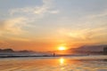 Sea with small waves, sand on the beach, mountains in the background, clouds from the sky, the sun sets, reflections in water and