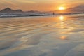 Sea with small waves, sand on the beach, mountains in the background, clouds from the sky, the sun sets, reflections in water and