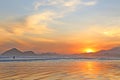 Sea with small waves, sand on the beach, mountains in the background, clouds from the sky, the sun sets, reflections in water and