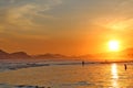 Sea with small waves, sand on the beach, mountains in the background, clouds from the sky, the sun sets, reflections in water and