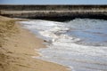 Seagull on deserted beach stands at water`s edge. Waves run on sandy beach. In background is pier, built of stone. Beach story.