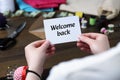 Seamstress holding paper note with phrase Welcome Back at table in atelier, closeup Royalty Free Stock Photo