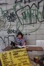 A seamstress working on the sidewalk in front of a wall covered with graffiti in a street of Bangkok