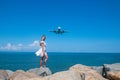 Seaside Fantasy: Girl in White Dress, Stones, Blue Sea, and a Flying Plane Royalty Free Stock Photo