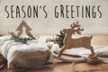 Season`s greetings text sign on stylish christmas rustic gift wrapped in linen fabric with green branch on wood with pine cones,