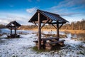 A seating area with a table, benches and a roof by the Venta River on a sunny, snowy autumn day. Kuldiga, Latvia