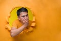 Young Asian man covering his mouth with hand, looking at a copy space, poses through torn yellow paper hole Royalty Free Stock Photo