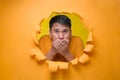 Secret, silence concept. Young Asian man covering his mouth with hand poses through torn yellow paper hole, wearing yellow t-shirt Royalty Free Stock Photo