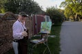 A cheerful elderly artist paints a picture with oil paints on a rural street.