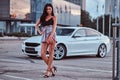 Seductive brunette woman dressed in modern clothes holds handbag posing near luxury car against a skyscraper. Royalty Free Stock Photo