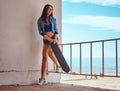 Seductive hot brunette girl wearing short shorts and jeans jacket holds a skateboard while standing to lean on a Royalty Free Stock Photo