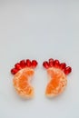 Segments of orange mandarin and pomegranate seeds looking like kid`s feet on white background. Vertical. Copy space. Royalty Free Stock Photo