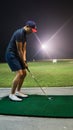 A man with a short pants swinging golf clubs at the driving range during the night