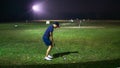 A man with a short pants holding a golf club playing at the golf driving range on a ground during the night