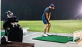 A man with a short pants holding a golf club playing at the golf driving range on a green carpet mat during the night