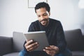 Selective focus.Attractive bearded African man reading news on digital tablet while sitting sofa in his modern studio Royalty Free Stock Photo