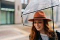 Selective focus of charming young woman wearing elegant hat standing with transparent umbrella on city street enjoying Royalty Free Stock Photo