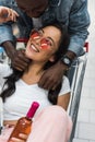 Focus of cheerful african american man hugging asian woman in sunglasses holding bottle and sitting in shopping cart Royalty Free Stock Photo