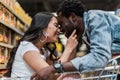 Focus of cheerful asian girl sitting in shopping cart and touching face of handsome and happy african american man Royalty Free Stock Photo