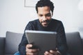 Selective focus.Closeup view of attractive bearded African man reading news on digital tablet while sitting sofa in his Royalty Free Stock Photo