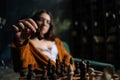 Selective focus of cute businesswoman in elegant eyeglasses making chess move sitting on armchair in dark room, on Royalty Free Stock Photo