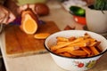 Enamel vintage bowl with sweet potato wedges against blurred chef cutting whole tubers of organic batata on wooden board
