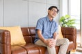 Selective focus at face of old senior Asian man sitting at sofa touching knee and legs suffer from the pain and injury.