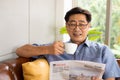 Selective focus at face. Senior Asian man reading newspaper mock up at the sofa couch while drinking coffee in the morning. Happy Royalty Free Stock Photo