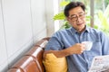 Selective focus at face. Senior Asian man reading newspaper mock up at the sofa couch while drinking coffee in the morning. Happy Royalty Free Stock Photo