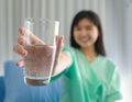 Selective focus on glass. Young inpatient woman who is getting well from her ailment smiling on to a camera and holding a glass