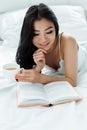 Focus of happy brunette asian woman reading book and holding cup of tea Royalty Free Stock Photo
