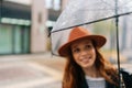 Selective focus of happy young woman in hat standing with transparent umbrella on city street enjoying rainy weather Royalty Free Stock Photo