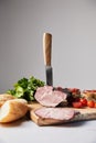 Focus of knife in tasty ham on cutting board with parsley, cherry tomatoes and baguette on white surface isolated on