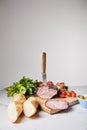 Focus of knife in tasty ham on cutting board with parsley, cherry tomatoes and baguette on white surface isolated on