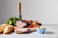 Focus of knife in tasty ham on cutting board with parsley, cherry tomatoes, olives and baguette on white surface