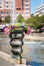 Selective focus of a large bronze vase with colorful flowers at a city square with water fountain and buildings.