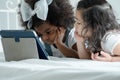 Selective focus on little African kid girl lying on white bed put hands on chin and using tablet with Caucasian cute friend Royalty Free Stock Photo